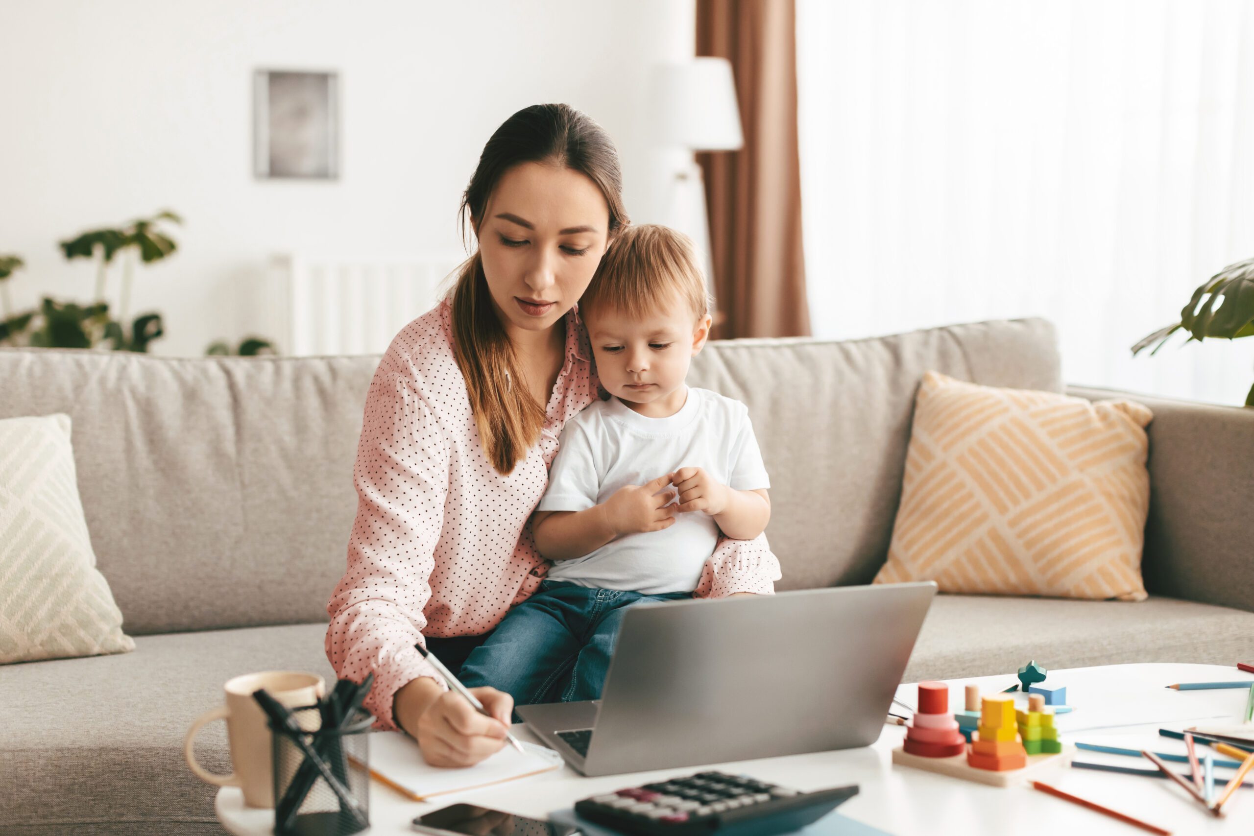 A mother reviewing court documents on a laptop at a coffee table, taking notes while holding her toddler on her lap.