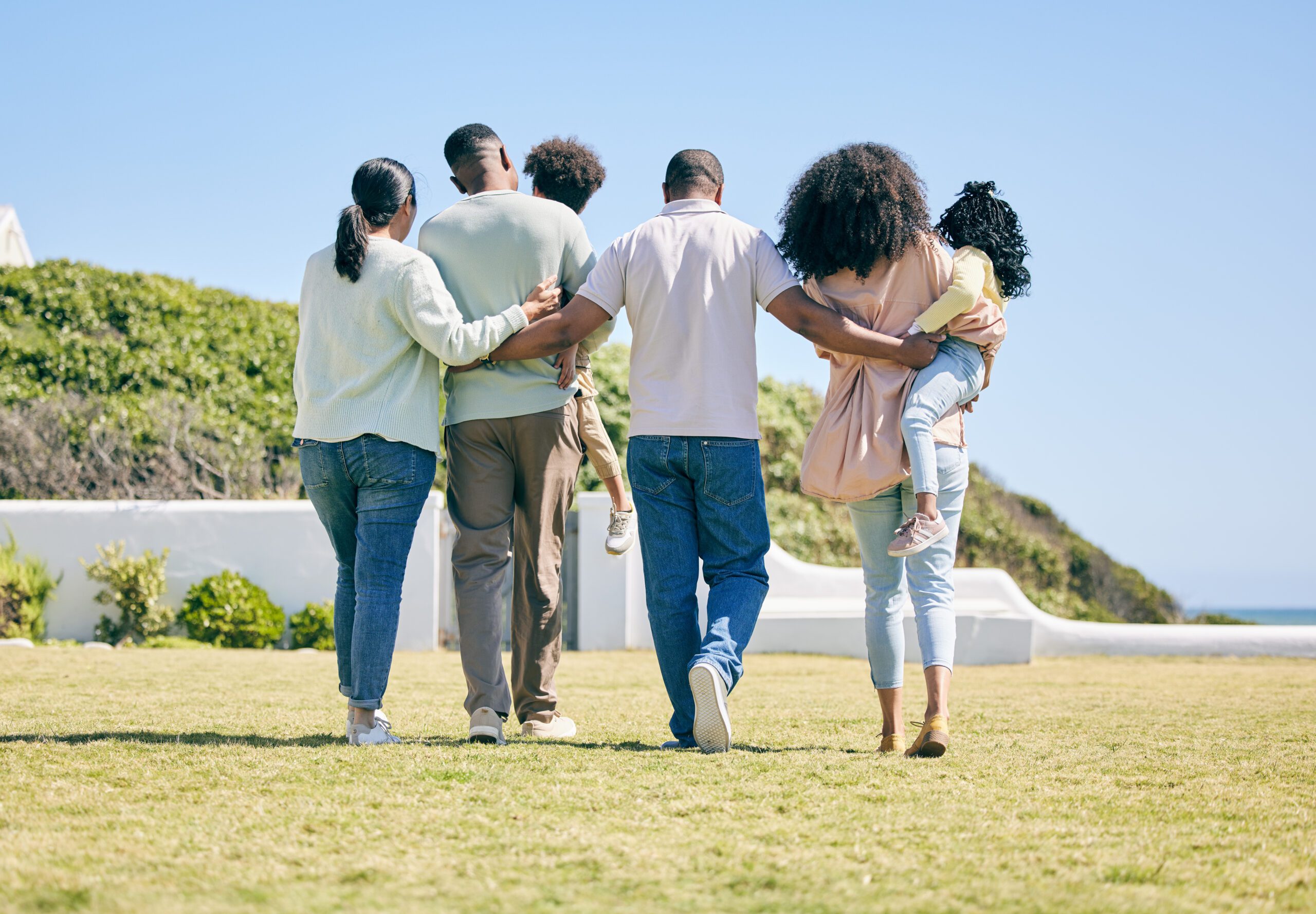 Back view of three generations of a family—grandparents, parents, and children—embracing as they walk away together through a garden.